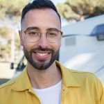 A cheerful bearded man wearing glasses is smiling outdoors in a natural setting.
