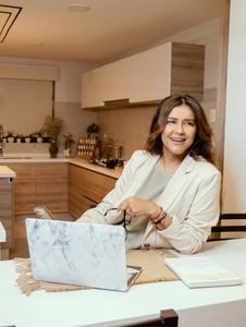 A woman in a cozy kitchen setting, smiling while working on a laptop and holding glasses.